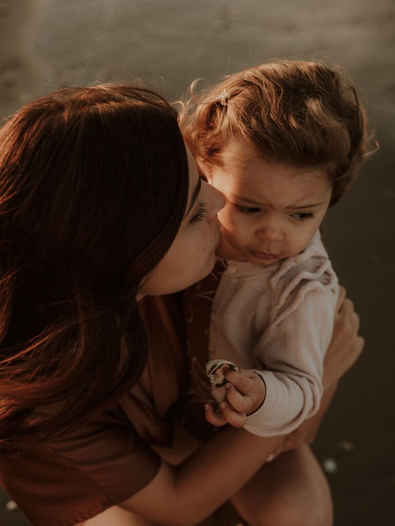 Mamma e bambina all’alba sulla spiaggia durante un servizio fotografico di famiglia, abbracciate in un momento intimo e dolce, con luce calda del mattino.